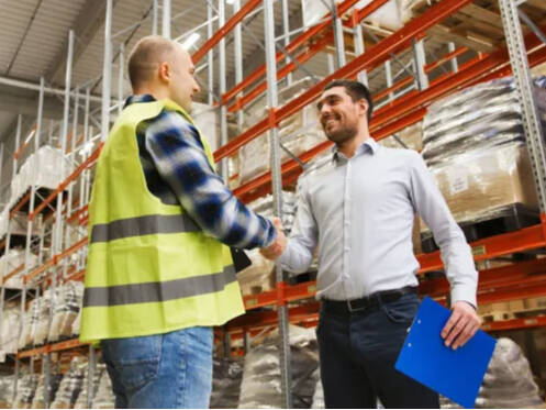 Two people shaking hands in a warehouse environment, one in a safety vest and one in a collared shirt and clipboard